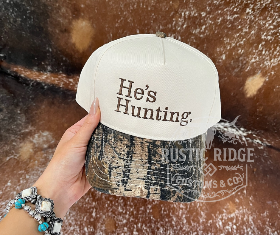He’s Hunting embroidered trucker hat with camo bill and neutral crown by Rustic Ridge Customs & Co