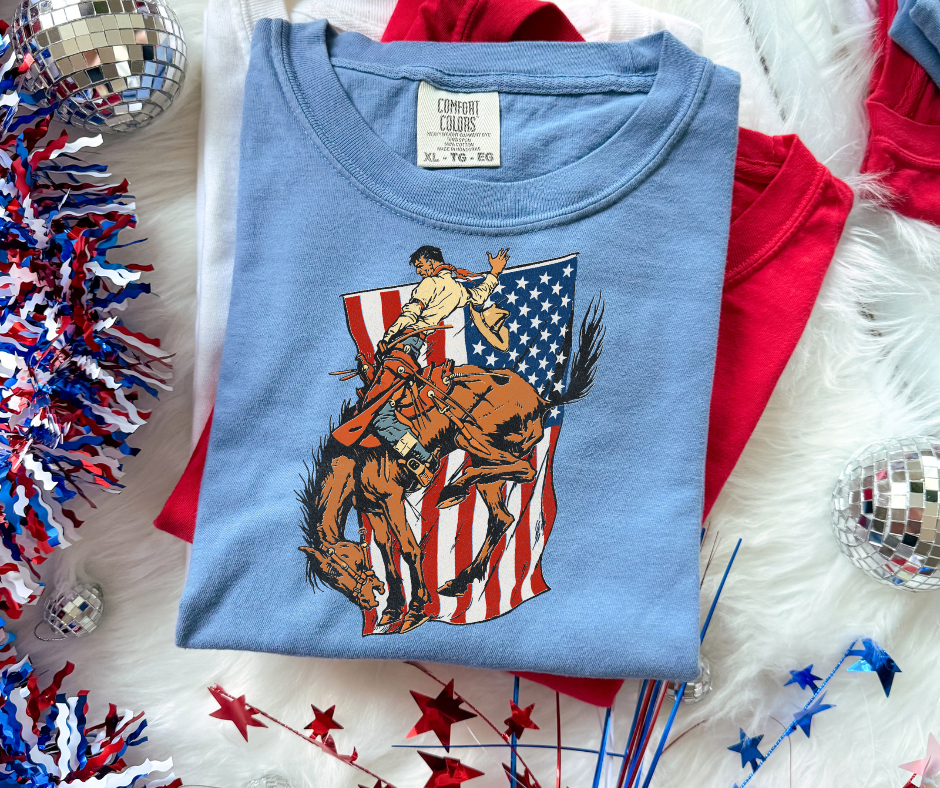 Patriotic western graphic tee featuring a vintage bronc rider and American flag design on a Comfort Colors shirt by Rustic Ridge Customs & Co.