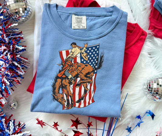 Patriotic western graphic tee featuring a vintage bronc rider and American flag design on a Comfort Colors shirt by Rustic Ridge Customs & Co.